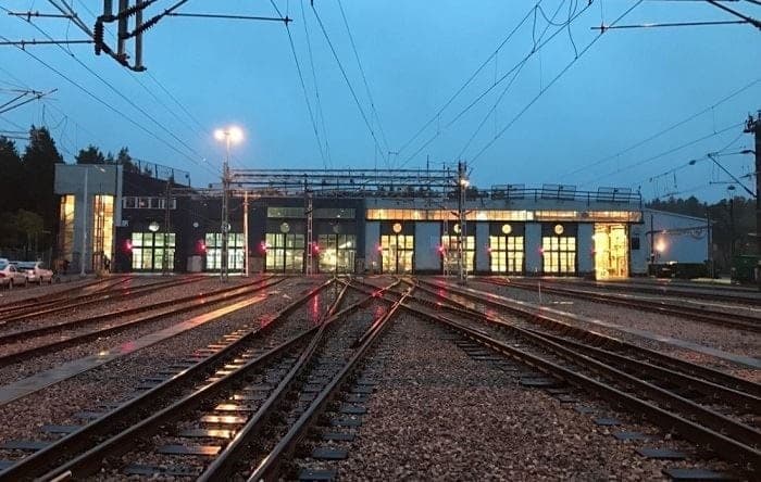 Spår och kontaktledningar leder fram till Älvsjödepåns upplysta depåhall i skymningen