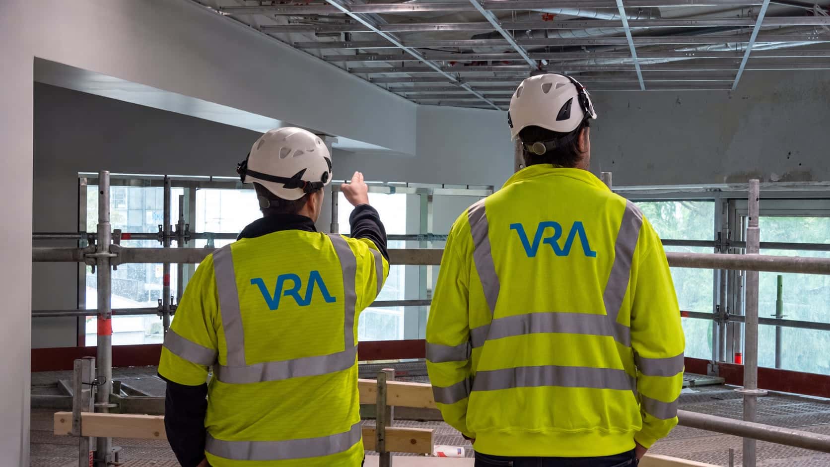 Två VRA-medarbetare i varseljackor och hjälm, sedda bakifrån på ett bygge, där den ena pekar mot fönstren