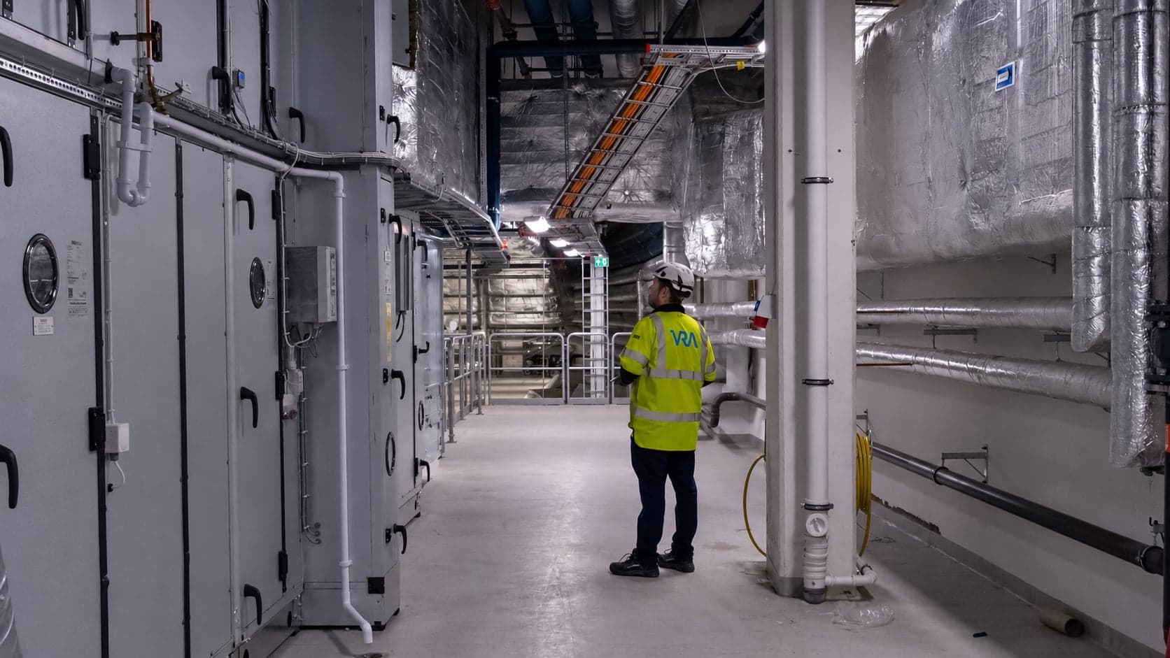 En VRA-medarbetare i varselkläder och hjälm i en lång korridor med ventilationsaggregat och isolerade kanaler