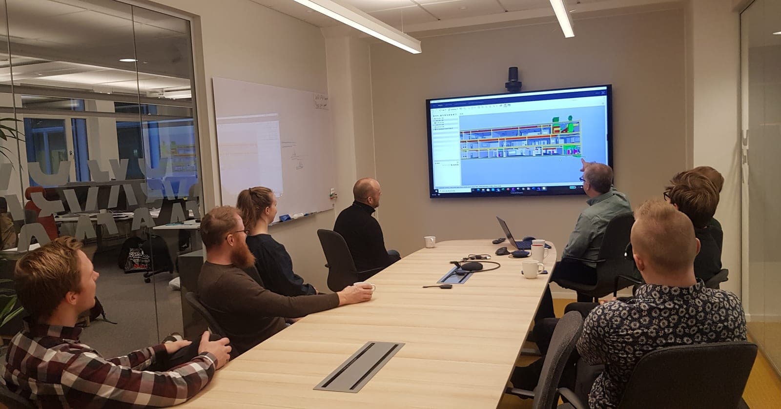 Mötesdeltagare runt ett konferensbord tittar på en byggnadsmodell som visas på en stor skärm